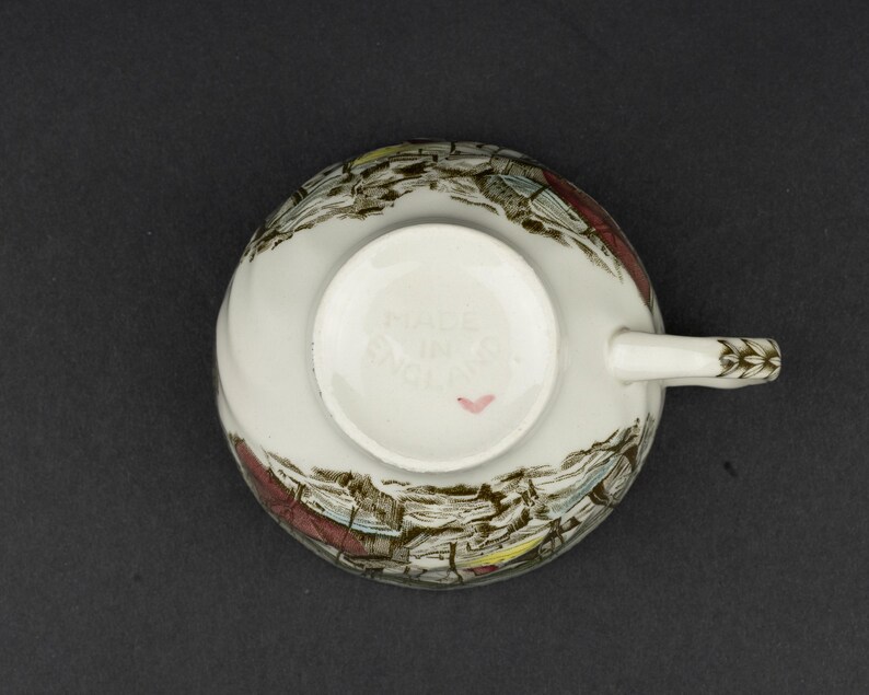 Pu&ograve; includere: Una tazza da t&egrave; in ceramica bianca vista dal basso, caratterizzata da una stampa paesaggistica in marrone e multicolore. La base presenta la scritta in rilievo MADE IN ENGLAND e un piccolo cuore rosa dipinto a mano vicino al bordo.
