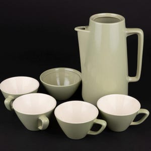 May include: A sage green ceramic coffee set consisting of a tall pitcher with a handle and spout, a small bowl, and four matching cups. Each piece features a smooth, light sage green exterior finish and a contrasting creamy white interior surface.