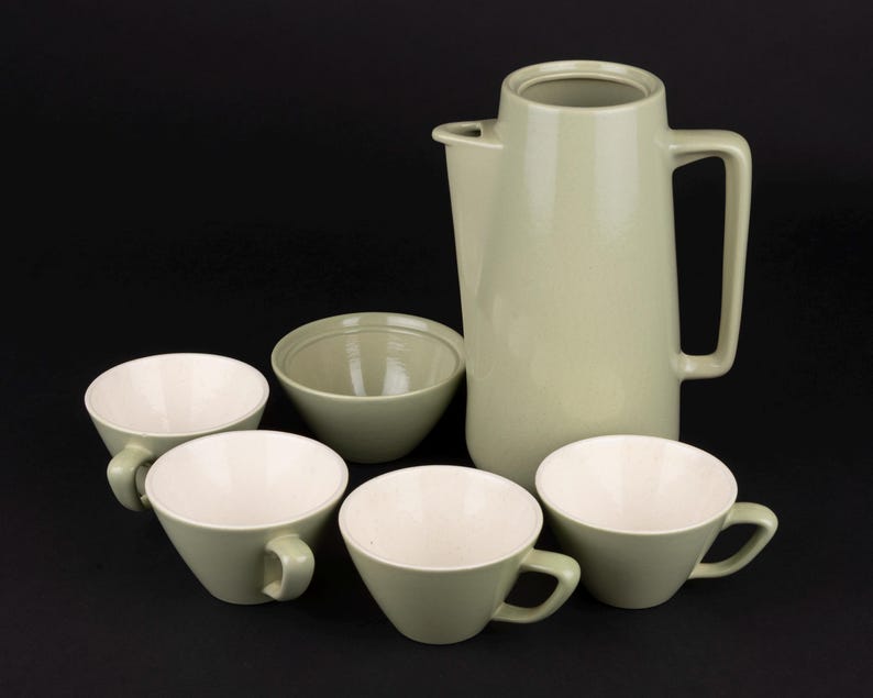 May include: A sage green ceramic coffee set consisting of a tall pitcher with a handle and spout, a small bowl, and four matching cups. Each piece features a smooth, light sage green exterior finish and a contrasting creamy white interior surface.