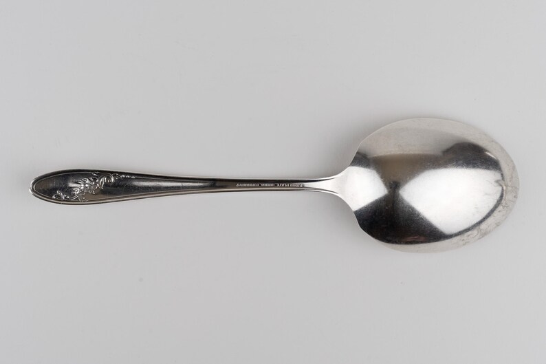 May include: A silver-toned metal spoon featuring an oval bowl and a slender handle. The handle is decorated with an embossed floral pattern near the tip and includes a small, stamped inscription along the center of the stem.