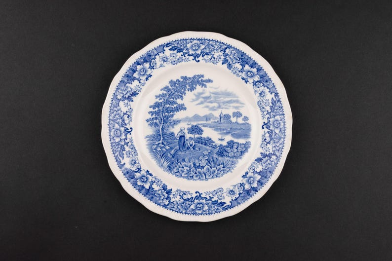 May include: A white ceramic plate with a scalloped edge featuring a blue transferware pattern. The center depicts a landscape with figures, trees, and a body of water, while the rim is decorated with a dense floral and leaf border design.