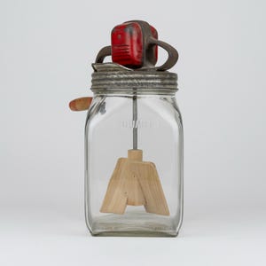 May include: A vintage glass butter churn with a square jar embossed with 2 QUARTS. It features a metal lid, a red painted gear housing, a hand crank with a wooden handle, and an internal wooden paddle mechanism for churning cream into butter.