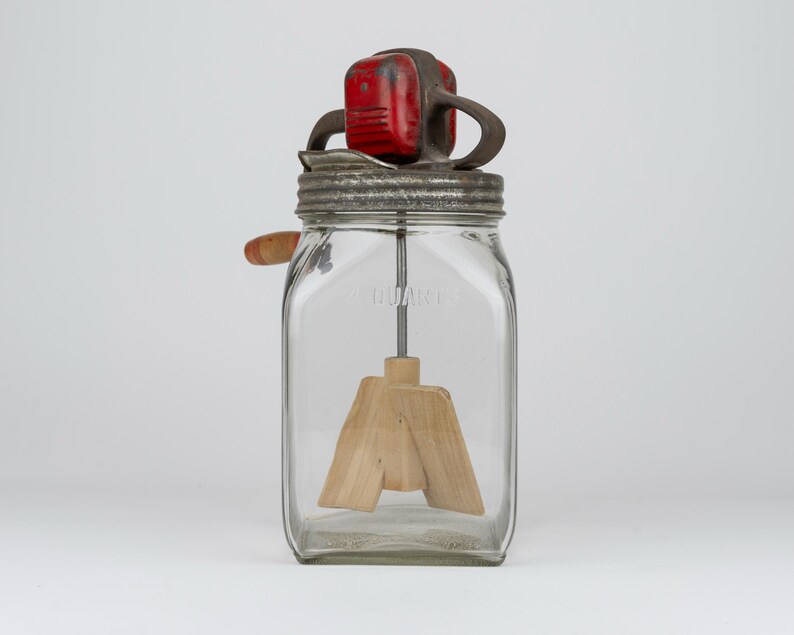 May include: A vintage glass butter churn with a square jar embossed with 2 QUARTS. It features a metal lid, a red painted gear housing, a hand crank with a wooden handle, and an internal wooden paddle mechanism for churning cream into butter.