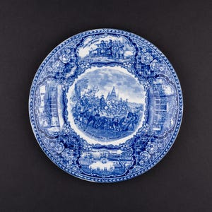 May include: A round ceramic plate featuring a blue and white transferware pattern. The center depicts a historical scene with figures, while the border contains smaller vignettes and floral motifs. Text on the plate includes "Landing of the Pilgrims" and "John's Discovery of the Mississippi".