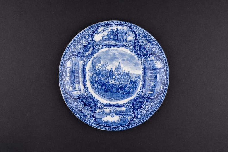 May include: A round ceramic plate featuring a blue and white transferware pattern. The center depicts a historical scene with figures, while the border contains smaller vignettes and floral motifs. Text on the plate includes "Landing of the Pilgrims" and "John's Discovery of the Mississippi".