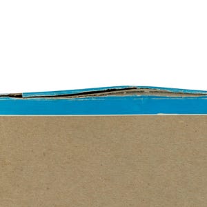 K&ouml;nnte beinhalten: Ein rechteckiger Pappkarton mit einer hellbraunen Mittelfl&auml;che und einem leuchtend blauen Rand. Die Oberkante des Kartons weist sichtbare Abnutzungserscheinungen auf, wobei sich die Pappschichten entlang der blauen Kante leicht abl&ouml;sen.