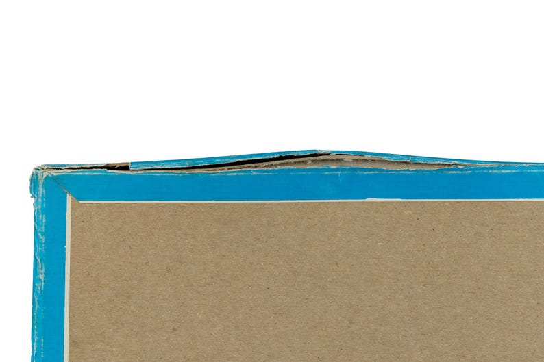 K&ouml;nnte beinhalten: Ein rechteckiger Pappkarton mit einer hellbraunen Mittelfl&auml;che und einem leuchtend blauen Rand. Die Oberkante des Kartons weist sichtbare Abnutzungserscheinungen auf, wobei sich die Pappschichten entlang der blauen Kante leicht abl&ouml;sen.
