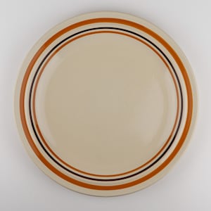 May include: A round ceramic plate with a smooth cream-colored center. The rim features concentric circular bands in shades of burnt orange and dark brown, creating a simple, clean design on the surface of the dinnerware piece.