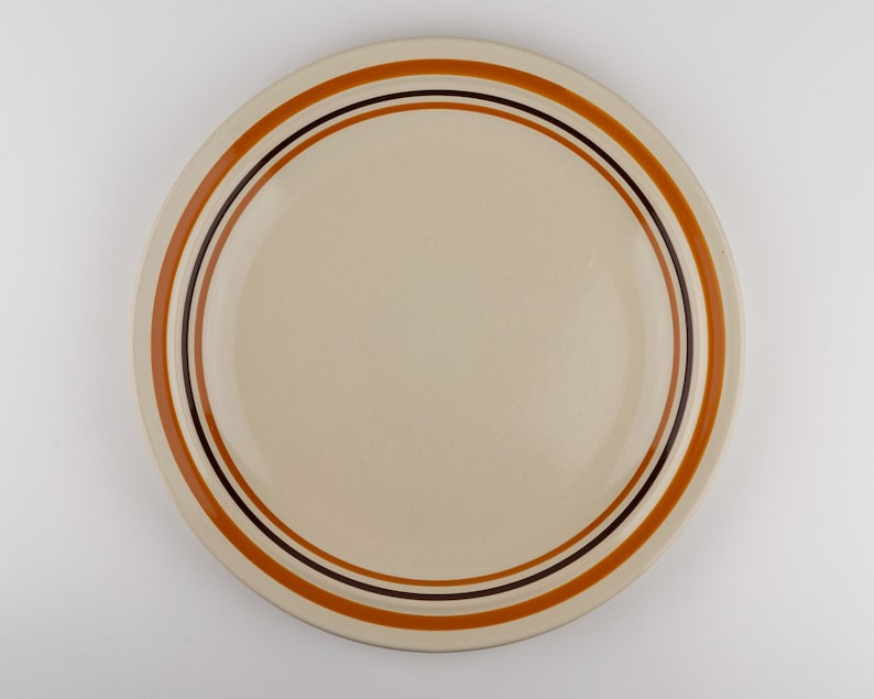 May include: A round ceramic plate with a smooth cream-colored center. The rim features concentric circular bands in shades of burnt orange and dark brown, creating a simple, clean design on the surface of the dinnerware piece.