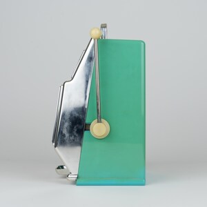 May include: A side view of a mint green slot machine featuring a polished chrome metal front panel and a vertical lever with a cream-colored knob. The device has a rectangular body with a flat base and a metallic finish on the front section.