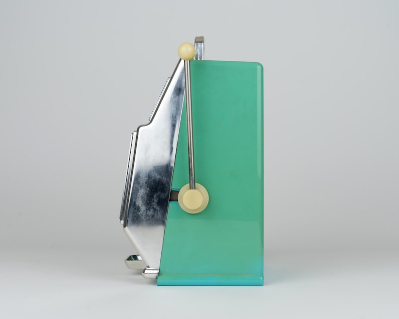 May include: A side view of a mint green slot machine featuring a polished chrome metal front panel and a vertical lever with a cream-colored knob. The device has a rectangular body with a flat base and a metallic finish on the front section.