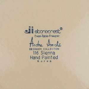 May include: A cream-colored ceramic plate base featuring blue printed text that reads: ji stonecrest Oven-Table-Freezer, Andre Ponche DESIGNER COLLECTION, 116 Sienna, Hand Painted, Korea. The surface is smooth with a circular rim indentation.