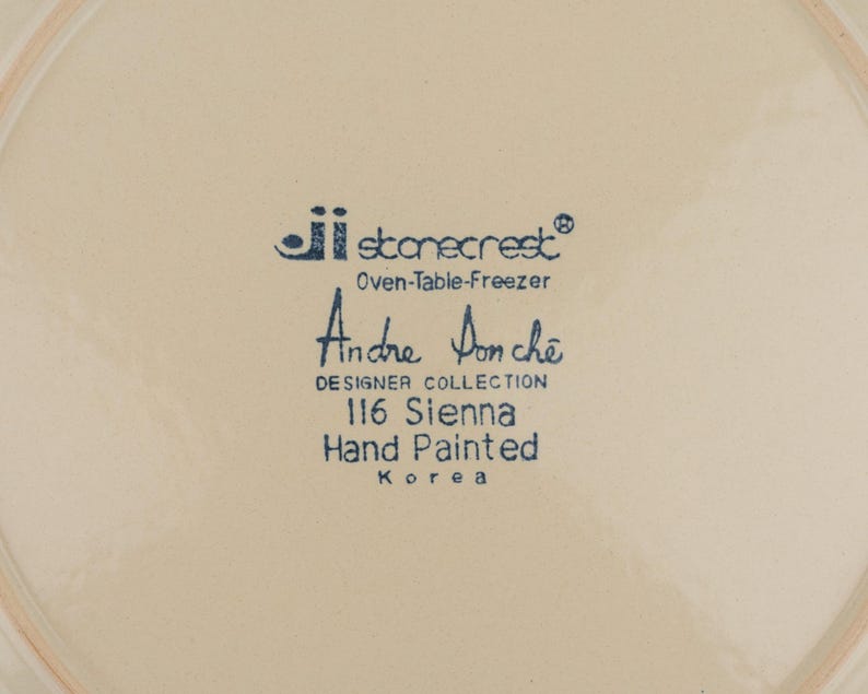 May include: A cream-colored ceramic plate base featuring blue printed text that reads: ji stonecrest Oven-Table-Freezer, Andre Ponche DESIGNER COLLECTION, 116 Sienna, Hand Painted, Korea. The surface is smooth with a circular rim indentation.