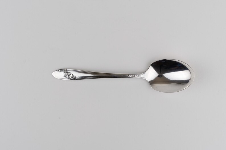 May include: A polished silver-tone metal spoon featuring a smooth, reflective bowl and a slender handle. The handle is decorated with a delicate, embossed floral pattern near the tip, adding a subtle texture to the otherwise sleek design.