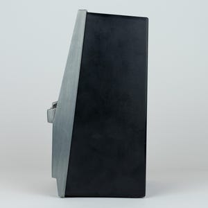 May include: A side profile view of a black rectangular electronic device featuring a brushed silver metallic front panel. The device has a tapered shape with a single silver control knob protruding from the side, set against a plain background.