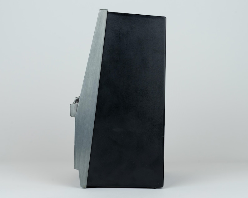 May include: A side profile view of a black rectangular electronic device featuring a brushed silver metallic front panel. The device has a tapered shape with a single silver control knob protruding from the side, set against a plain background.