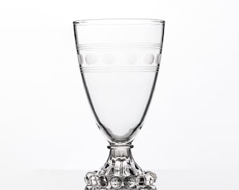 Puede incluir: Una copa de cristal transparente con un cuerpo liso y cónico, decorada con una banda horizontal de puntos y líneas grabadas. El tallo y la base presentan un patrón decorativo de cuentas texturizadas que contrasta con el cuerpo liso.