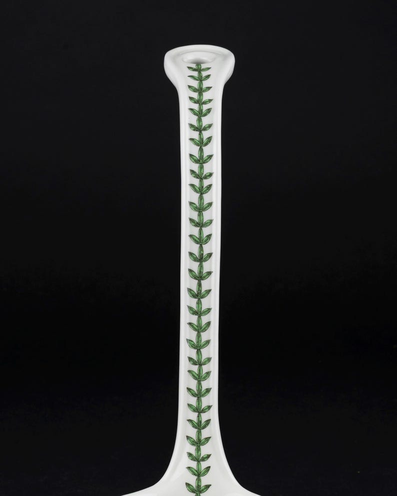 May include: A white ceramic candlestick holder featuring a slender, elongated neck. The front surface is decorated with a vertical, repeating pattern of small, detailed green leaves arranged in a symmetrical vine-like design along the center.