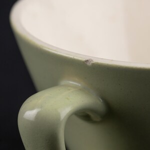 May include: A sage green ceramic mug with a smooth, curved handle and a cream-coloured interior. The upper rim of the mug features a small, visible chip in the glaze, revealing a light brown texture beneath the surface of the sage green exterior.