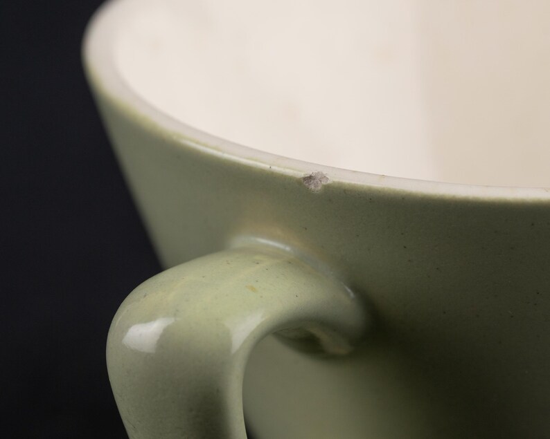 May include: A sage green ceramic mug with a smooth, curved handle and a cream-coloured interior. The upper rim of the mug features a small, visible chip in the glaze, revealing a light brown texture beneath the surface of the sage green exterior.