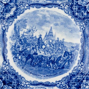 May include: A round ceramic plate featuring a blue and white transferware pattern. The center depicts a historical scene titled De Soto's Discovery of the Mississippi 1541. The border is decorated with floral motifs and architectural vignettes.