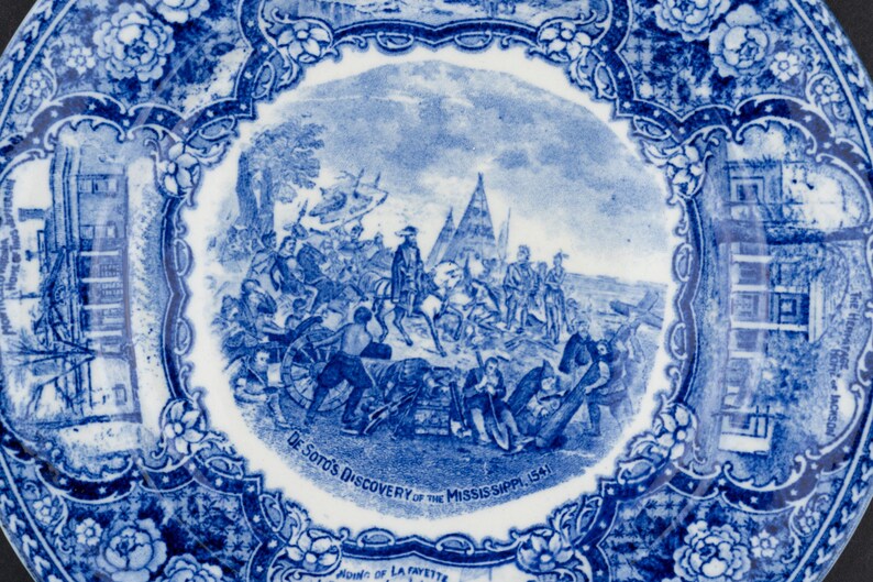 May include: A round ceramic plate featuring a blue and white transferware pattern. The center depicts a historical scene titled De Soto's Discovery of the Mississippi 1541. The border is decorated with floral motifs and architectural vignettes.