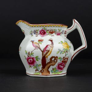 K&ouml;nnte beinhalten: Ein wei&szlig;es Sahnek&auml;nnchen aus Keramik mit einem handbemalten Motiv eines Fasans auf einem Ast, umgeben von leuchtend rosa, gelben und orangefarbenen Blumen, mit einem dekorativen gelb-schwarzen Rand entlang der oberen Kante des Gef&auml;&szlig;es.