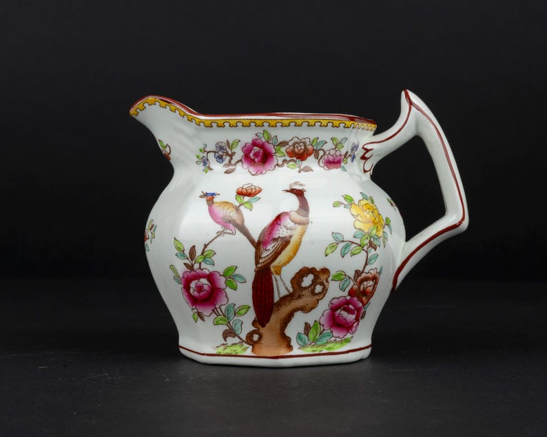 K&ouml;nnte beinhalten: Ein wei&szlig;es Sahnek&auml;nnchen aus Keramik mit einem handbemalten Motiv eines Fasans auf einem Ast, umgeben von leuchtend rosa, gelben und orangefarbenen Blumen, mit einem dekorativen gelb-schwarzen Rand entlang der oberen Kante des Gef&auml;&szlig;es.