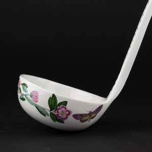 May include: A white ceramic soup ladle featuring a botanical illustration of pink flowers with green leaves and a purple moth on the exterior bowl. The long handle extends upward, displaying a matching pattern of small black and white floral motifs.