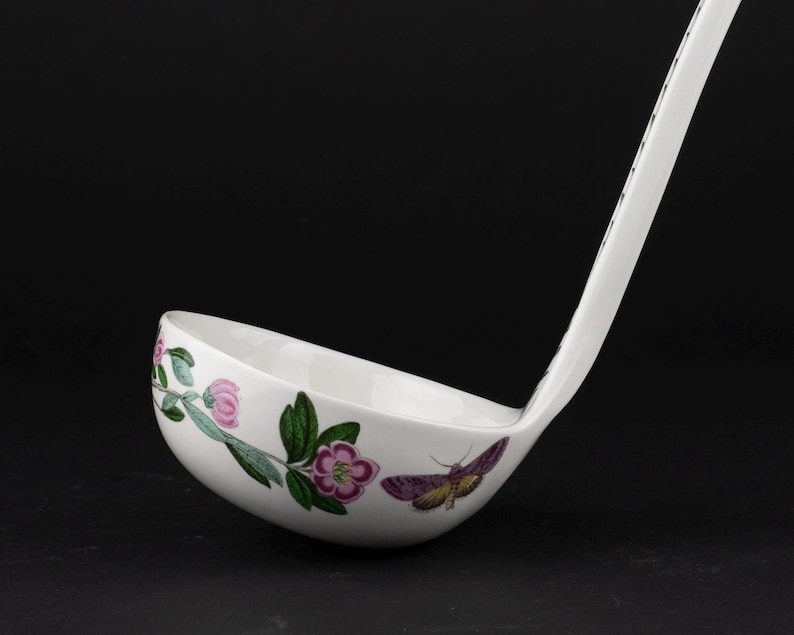 May include: A white ceramic soup ladle featuring a botanical illustration of pink flowers with green leaves and a purple moth on the exterior bowl. The long handle extends upward, displaying a matching pattern of small black and white floral motifs.