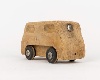 May include: A handcrafted wooden toy vehicle with a natural light brown finish. The bus-shaped body features carved window outlines, a single silver metal circular accent on the front, and four black rubber wheels with silver metal hubs.