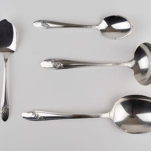 May include: A set of four silver-toned metal utensils including a small spoon with a chipped edge, a standard spoon, a small ladle, and a large serving spoon. Each handle features a delicate embossed floral pattern along the lower section.