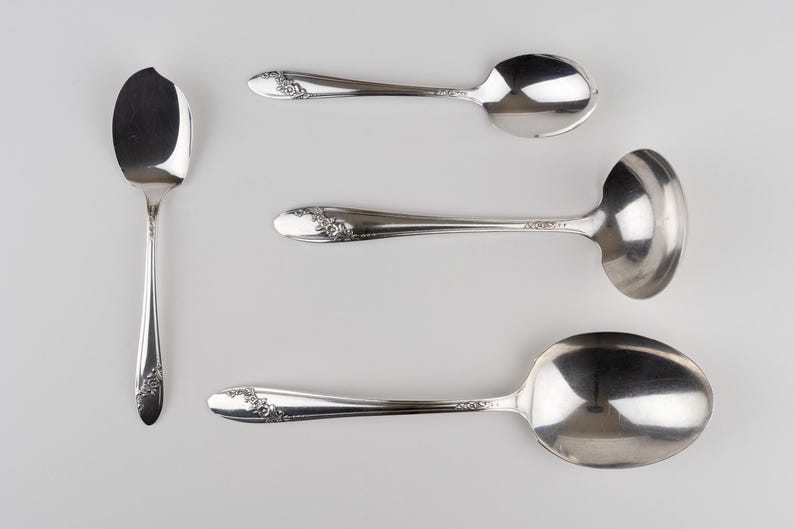 May include: A set of four silver-toned metal utensils including a small spoon with a chipped edge, a standard spoon, a small ladle, and a large serving spoon. Each handle features a delicate embossed floral pattern along the lower section.