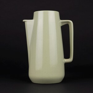 May include: A pale sage green pitcher with a smooth, matte finish. The vessel features a tapered cylindrical body, a wide integrated spout on the left side, and a large, angular handle attached to the right side, creating a clean, uniform look.