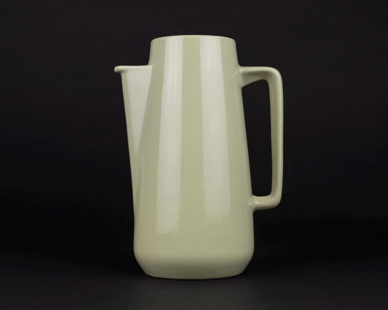 May include: A pale sage green pitcher with a smooth, matte finish. The vessel features a tapered cylindrical body, a wide integrated spout on the left side, and a large, angular handle attached to the right side, creating a clean, uniform look.