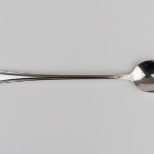 May include: A silver-toned metal spoon featuring a long, slender handle with an embossed floral pattern at the tip and a smaller decorative detail near the bowl. The spoon has a polished, reflective surface and a smooth, rounded oval bowl.