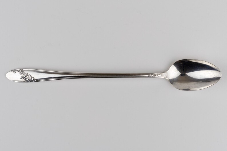 May include: A silver-toned metal spoon featuring a long, slender handle with an embossed floral pattern at the tip and a smaller decorative detail near the bowl. The spoon has a polished, reflective surface and a smooth, rounded oval bowl.