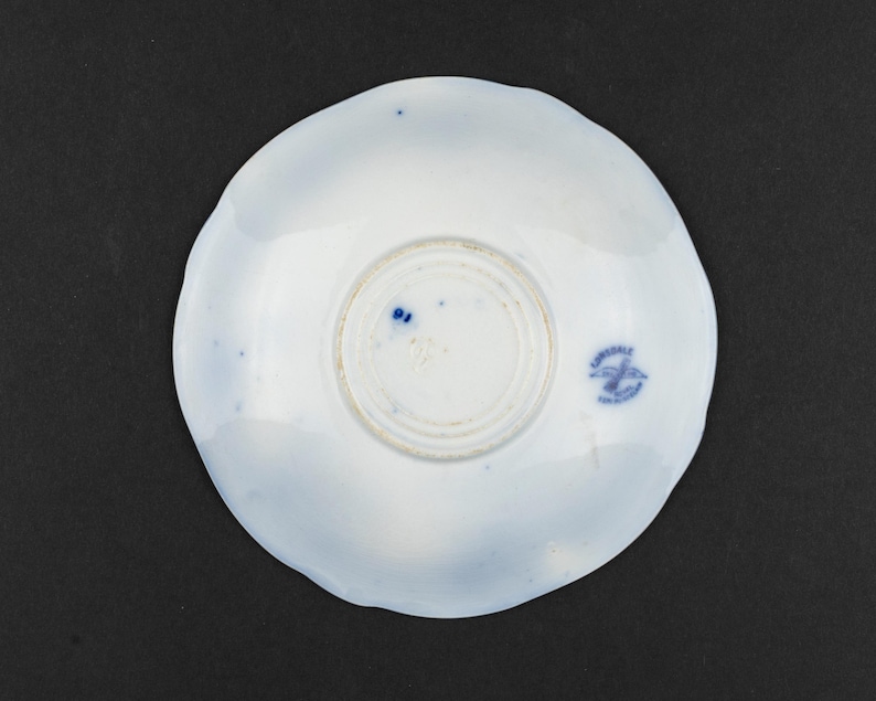 Puede incluir: Un platillo de cer&aacute;mica blanca con borde festoneado y una marca de fabricante impresa en azul en la parte inferior. La marca presenta un emblema de ave y el texto Lonsdale England. La superficie tiene sutiles manchas azules y un pie.