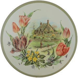 Może przedstawiać: Okrągły talerz ceramiczny z centralną ilustracją kamiennego domku z dachem krytym strzechą i ścieżką ogrodową. Scena jest otoczona kolorową kompozycją r&oacute;żowych i czerwonych tulipan&oacute;w, ż&oacute;łtych pierwiosnk&oacute;w oraz małych fioletowych fiołk&oacute;w.