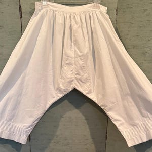May include: White bloomers, a vintage style garment, with a gathered waist and wide legs. The fabric is likely cotton or a similar lightweight material. The hem of each leg has a decorative, horizontal stitched detail.