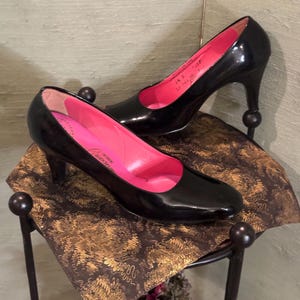 May include: A pair of black patent leather pumps with pink insoles and a stiletto heel. The shoes are on a gold and brown patterned surface.