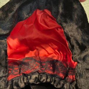 May include: A black faux fur hood with a vibrant red satin lining. The hood features a black lace trim along the bottom edge. The hood is designed to be worn as a costume accessory.