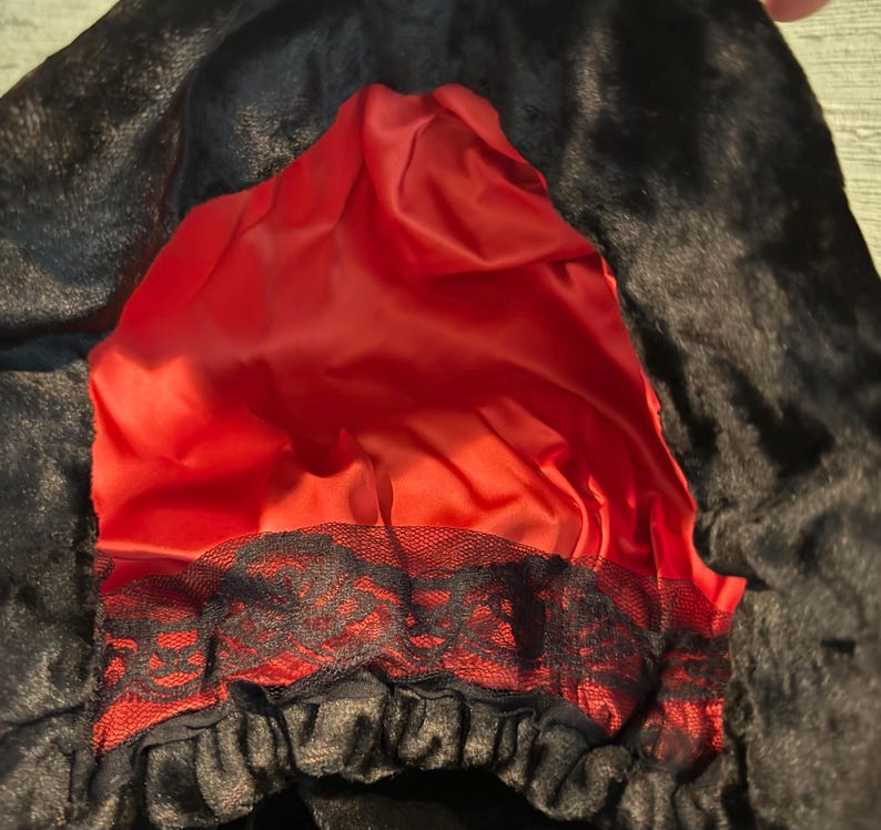 May include: A black faux fur hood with a vibrant red satin lining. The hood features a black lace trim along the bottom edge. The hood is designed to be worn as a costume accessory.