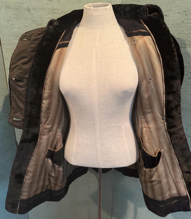 May include: A vintage black coat with a faux fur collar and lining, displayed on a mannequin. The coat features a tailored design with a button closure and two front pockets. The interior lining is a light brown color.
