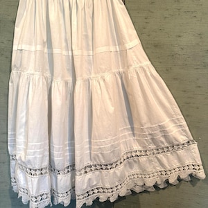 Puede incluir: Falda blanca escalonada con dobladillo de encaje festoneado. La falda está hecha de una tela ligera y cuelga de una cintura sencilla. La falda está suspendida de una percha.