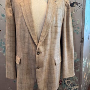 May include: A tan blazer with a subtle plaid pattern, featuring two front pockets and a notched lapel. The blazer is buttoned and worn over a white t-shirt and cream-colored pants. The blazer is displayed on a mannequin.