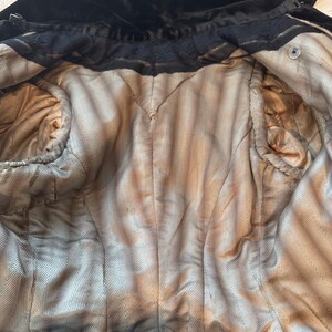 May include: An interior view of a vintage black velvet coat. The coat's lining is a light tan color with a herringbone pattern. The coat has a high collar and a button closure. The fabric appears to be aged.