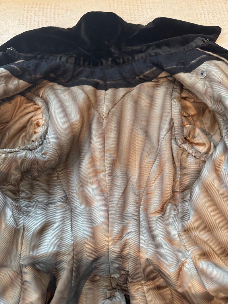 May include: An interior view of a vintage black velvet coat. The coat's lining is a light tan color with a herringbone pattern. The coat has a high collar and a button closure. The fabric appears to be aged.