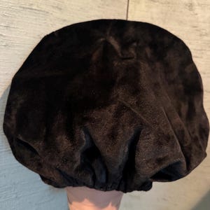 May include: A black velvet bonnet, likely for hair protection, is displayed. The bonnet is gathered at the base, creating a rounded shape. The fabric has a soft, textured appearance. The bonnet is placed on a mannequin head.