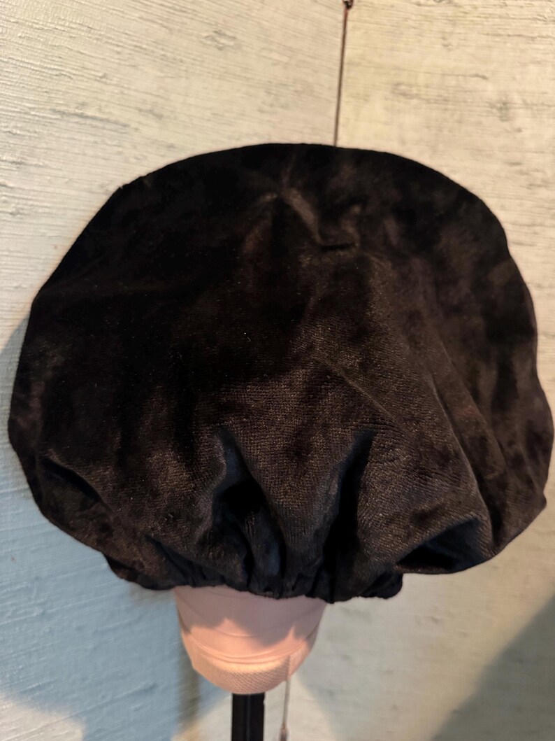 May include: A black velvet bonnet, likely for hair protection, is displayed. The bonnet is gathered at the base, creating a rounded shape. The fabric has a soft, textured appearance. The bonnet is placed on a mannequin head.